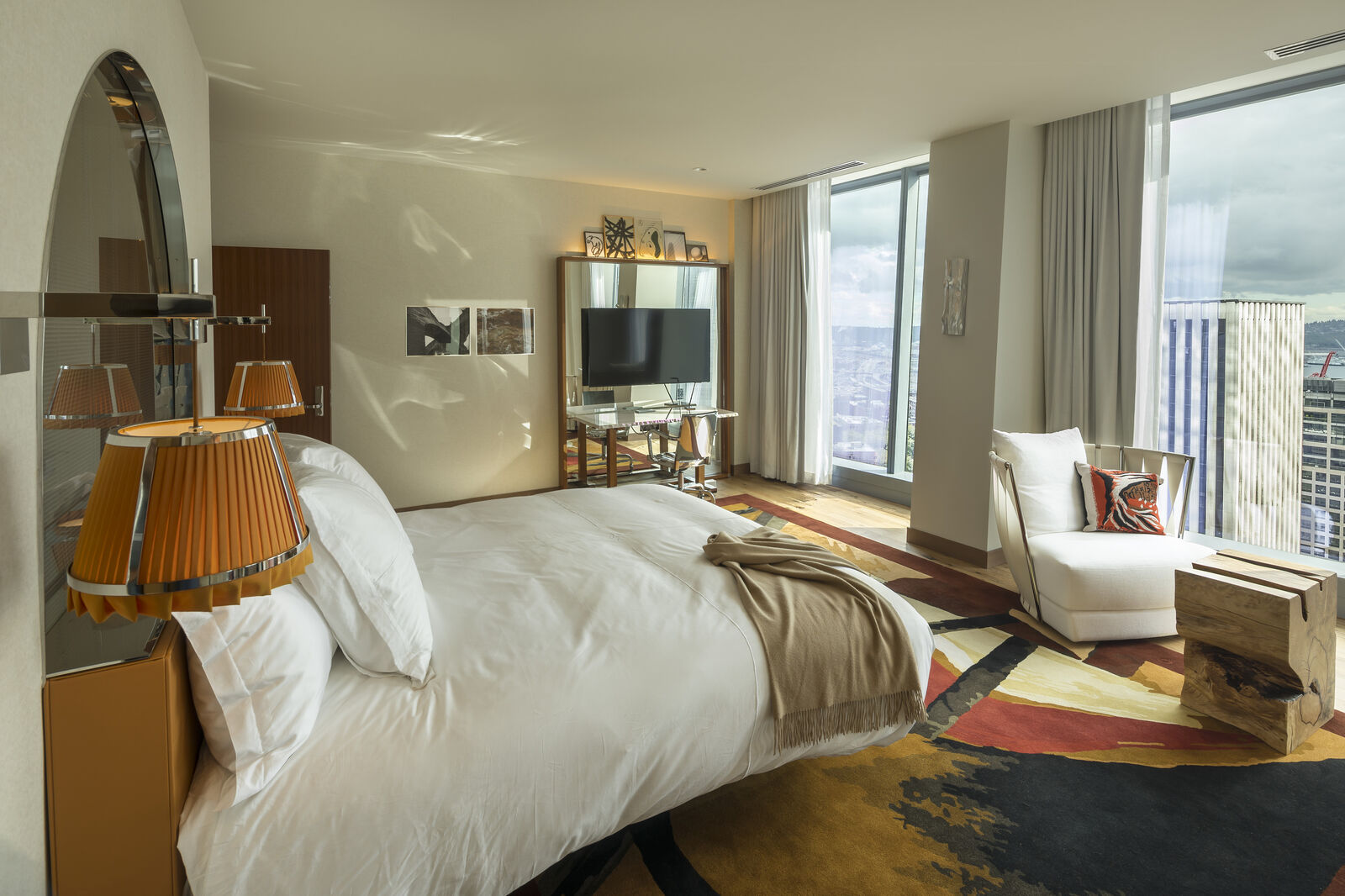 Interior photo of a hotel room in F5 Tower, showing a bed, table, chair, vanity, and full height windows looking out to the view of downtown Seattle